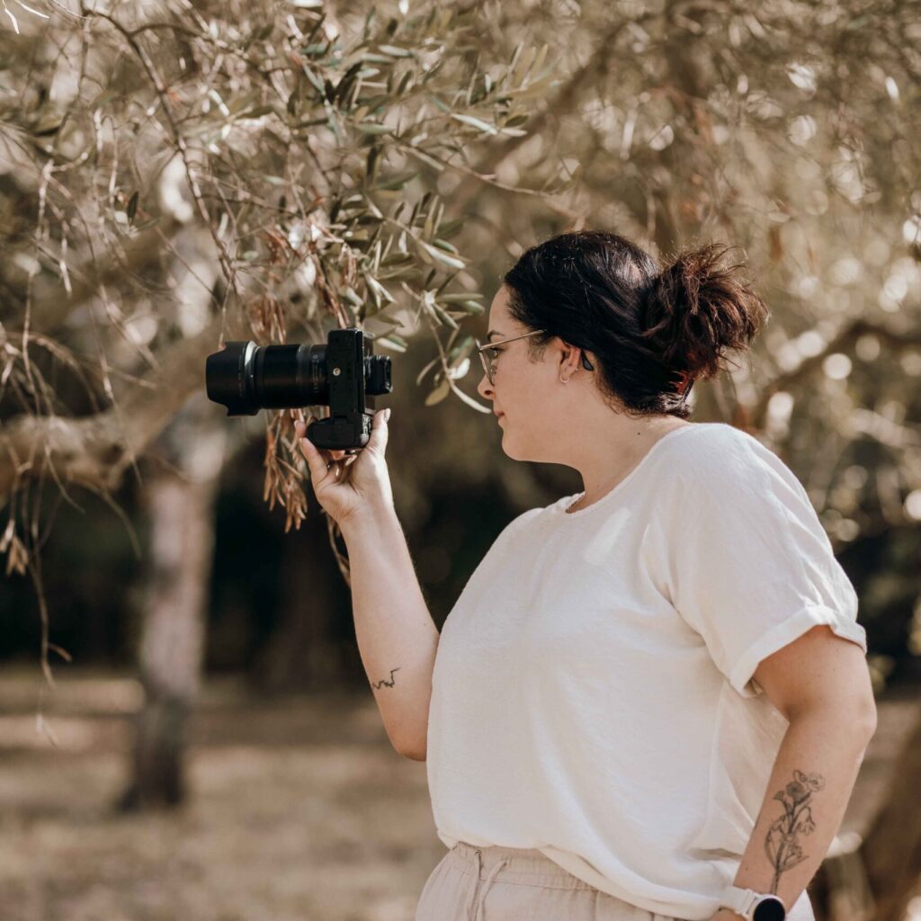 Eine Fotografin hält ihre Kamera hoch, während sie einen Olivenbaum fotografiert. Der Hintergrund ist voller Lichtreflexionen durch die Blätter.